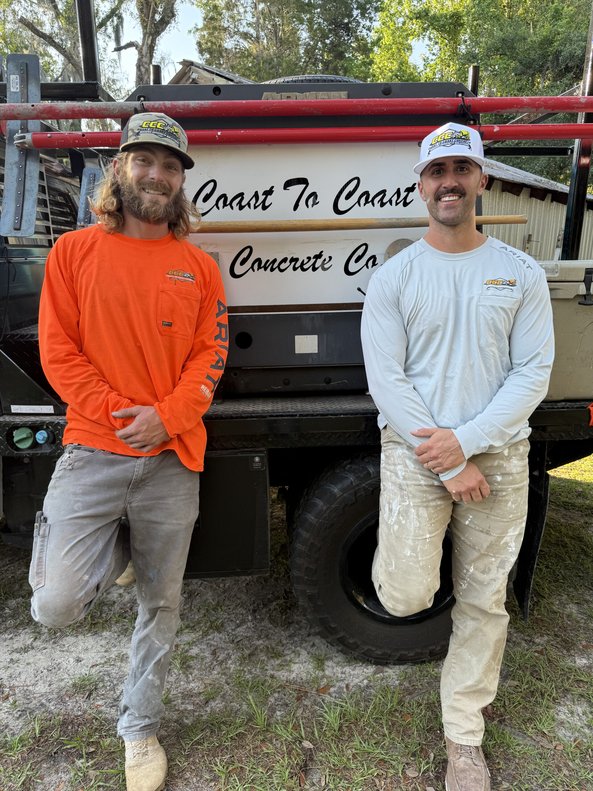 Two men in work clothes stand in front of a truck labeled Coast To Coast Concrete Coatings with equipment and trees in the background