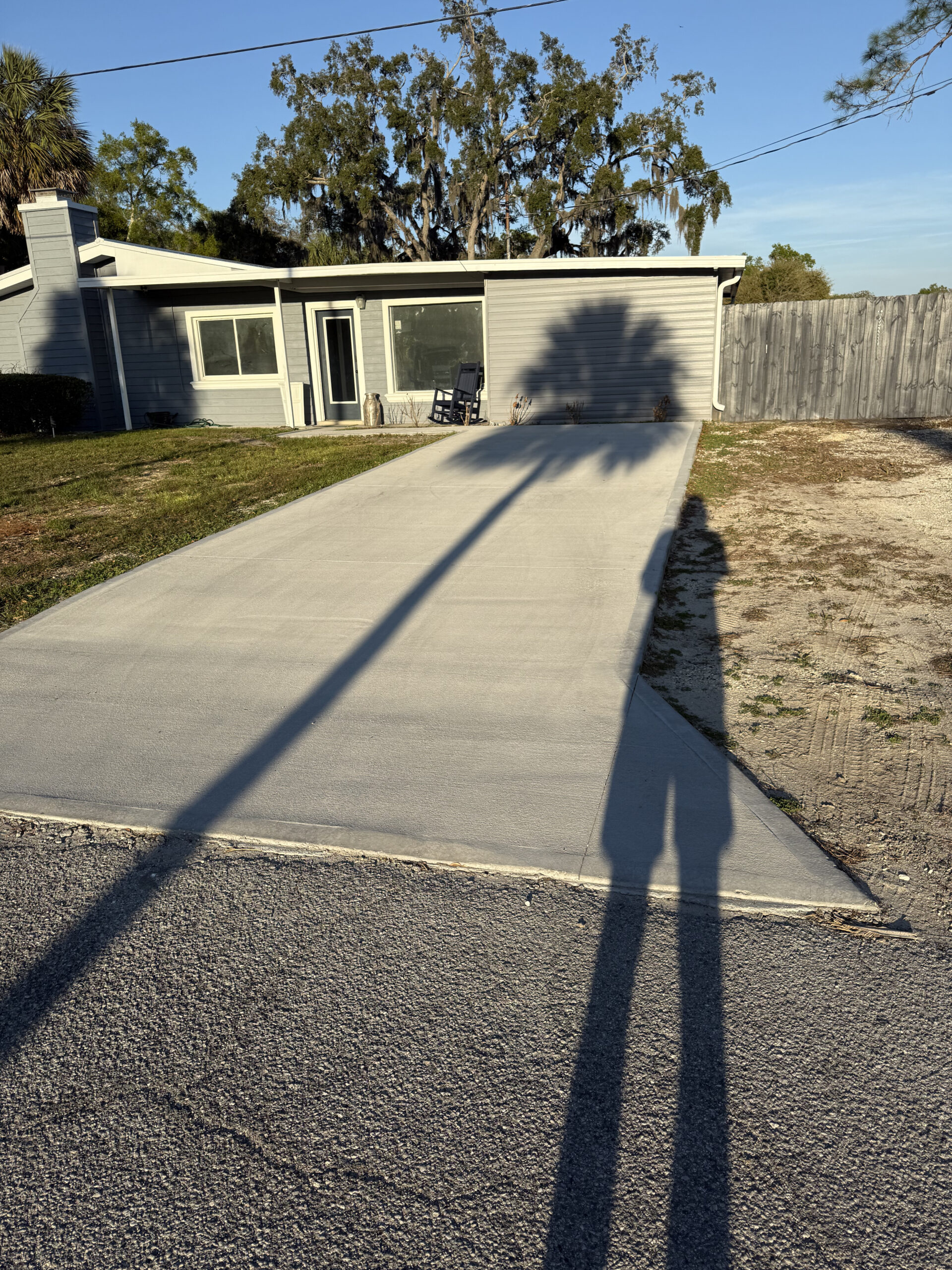 A concrete driveway leads to a one-story house with large windows; two tall palm tree shadows stretch across the driveway.
