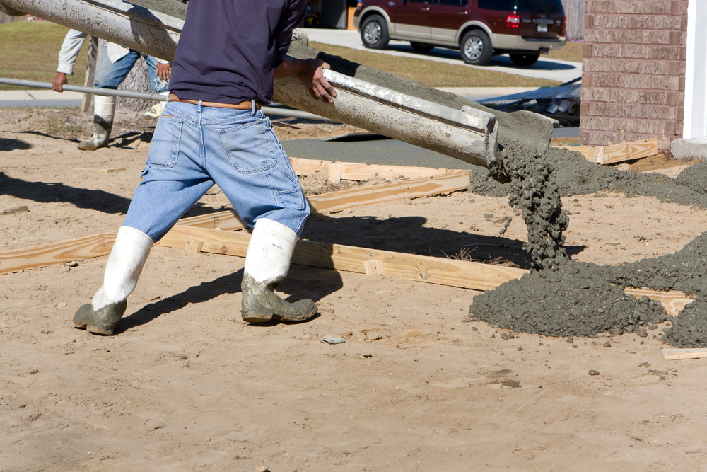 Worker in jeans and white boots pouring wet concrete from a chute onto a prepared foundation area at a construction site.