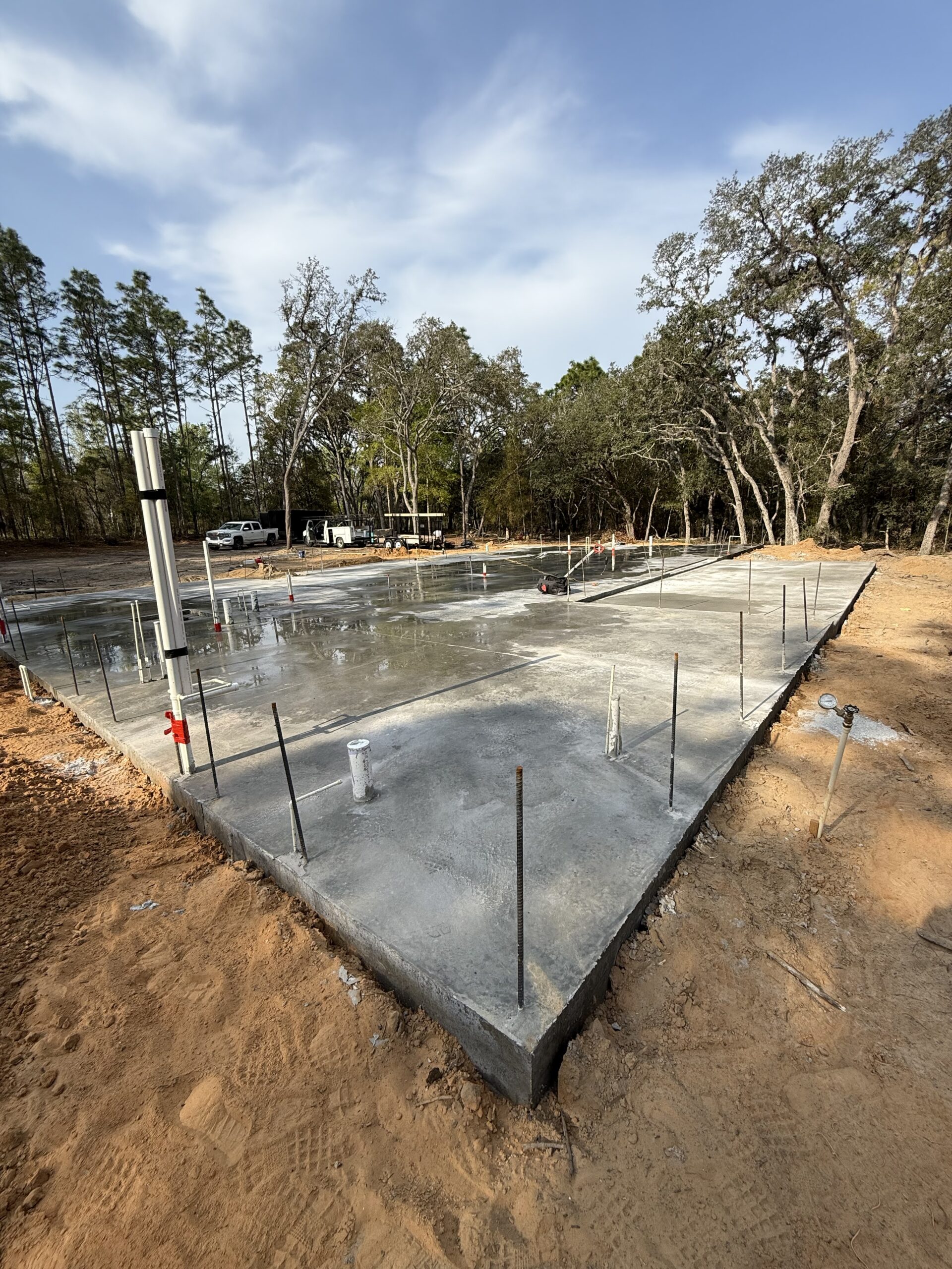Freshly poured concrete foundation slab with exposed plumbing and rebar, surrounded by dirt and trees, under a clear sky.