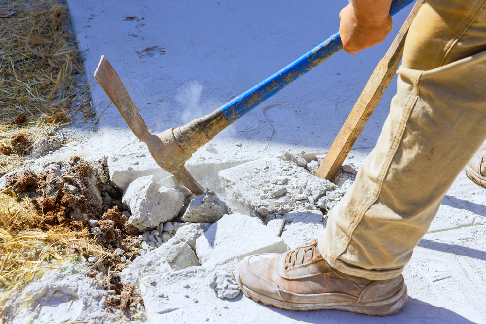 A person uses a pickaxe to break apart concrete on the ground, with debris and dust visible around the area.