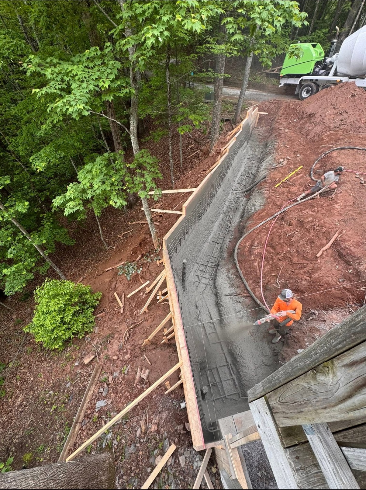 Two workers spray concrete onto a rebar-reinforced retaining wall beside a dirt slope, with formwork and a concrete mixer truck nearby in a wooded area.