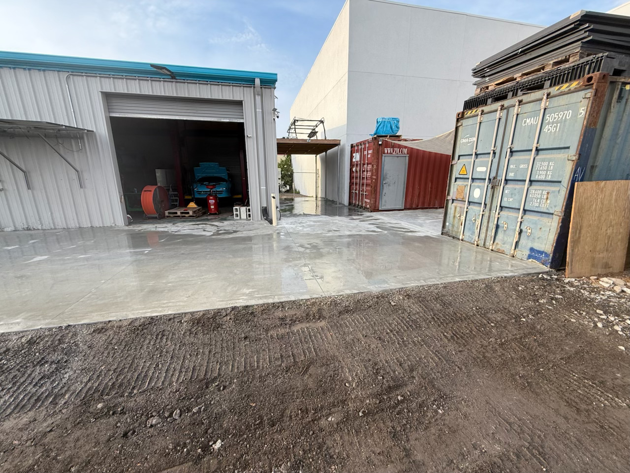 Industrial yard with a concrete surface, metal warehouse, stacked pallets, shipping containers, and equipment under a partly cloudy sky.