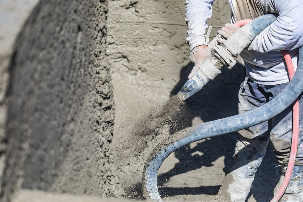 A construction worker provides shotcrete services, spraying concrete onto a large vertical surface with a hose, while concrete dust and debris fill the air.